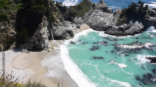 View of turquoise water at Big Sur State Park along the coast of California.  A waterfall can be seen through the rocks as well as algae in the blue-green ocean water.