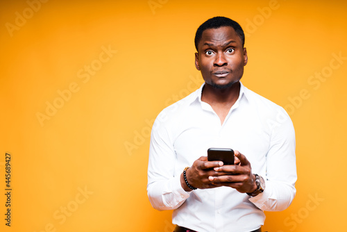 Handsome surprised african american man holding phone while standing on yellow background © DmitryStock