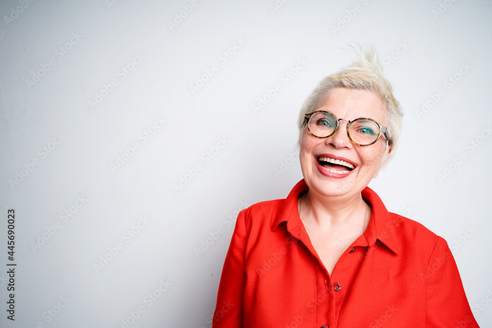 Happy elderly woman in shirt and glasses laughs opening mouth and squirts on gray background Happy elderly woman in shirt and glasses laughs opening mouth and squirts on gray background