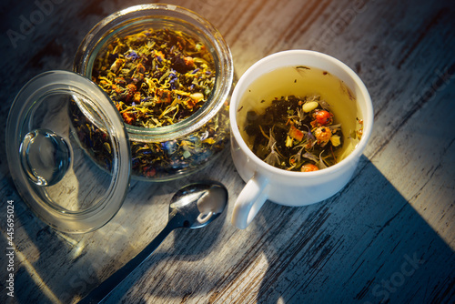 Dry mix of tea leaves, herbs, petals, berries and nuts in glass bowl. Natural herbal tea in white cup on gray wooden background in the evening light, shadow on the table.