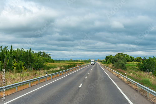 Landscape of a road lined with trees, cultivated fields and a cloudy sky, with a truck driving straight ahead.