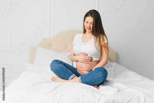 Portrait of nice adorable lovely sweet tender beautiful cheerful joyful pregnant curly-haired mom in casual wear sitting on divan, holding belly, white light interior room