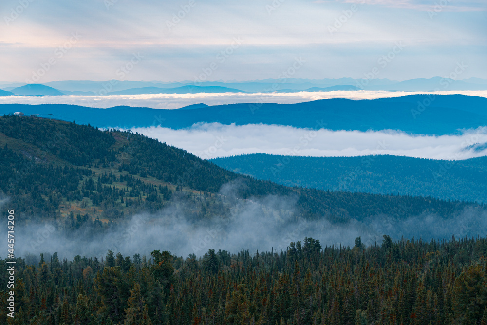 Panorama of a summer mountain valley with  peaks of the mountain range, clouds and fog. Forest in the foreground. View from the top.