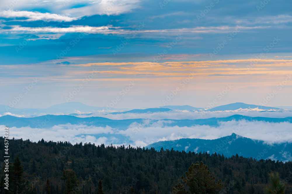Obraz premium Panorama of a summer mountain valley with peaks of the mountain range, clouds and fog. Forest in the foreground. View from the top.