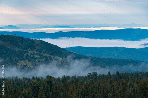 Panorama of a summer mountain valley with peaks of the mountain range, clouds and fog. Forest in the foreground. View from the top.