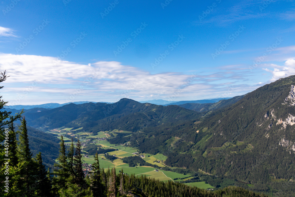 A panoramic view on the Alpine valley in Austria. High mountains around. The slopes are partially overgrown with small bushes, higher parts baren. Small lake at the bottom. Clear and sunny day.