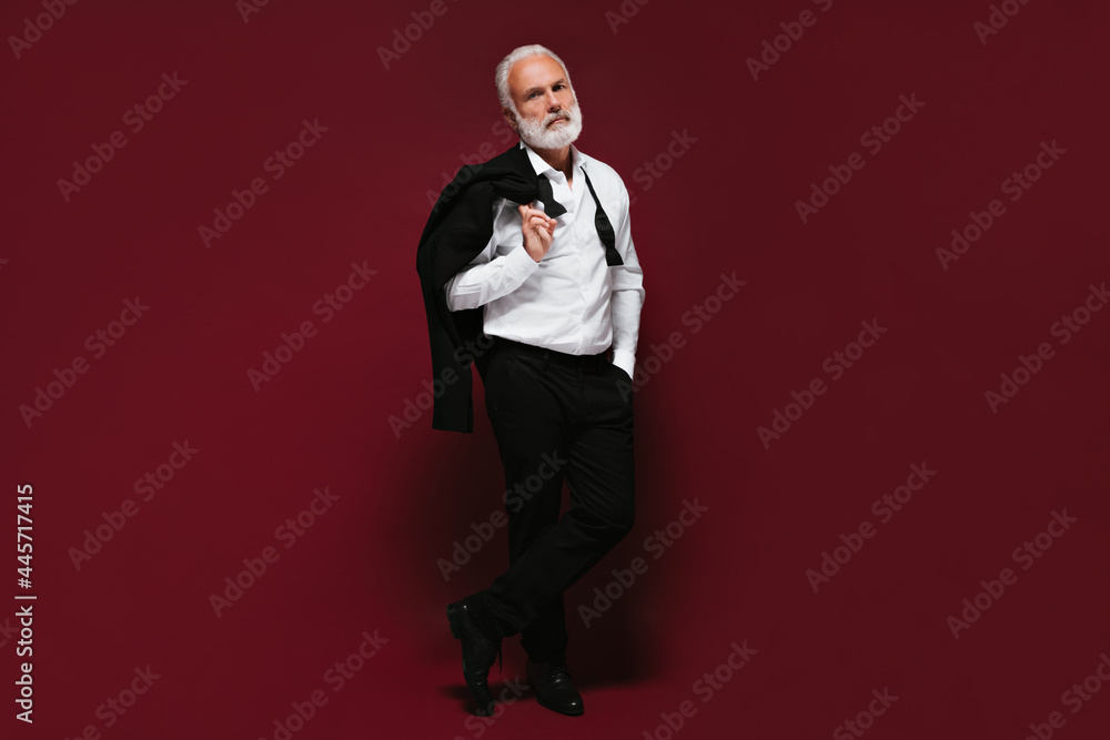 Serious man in suit looking into camera on burgundy background. Full length shot of guy with stylish beard in black pants and jacket.. Serious man in suit looking into camera on burgundy background. Full length shot of guy with stylish beard in black pants and jacket..