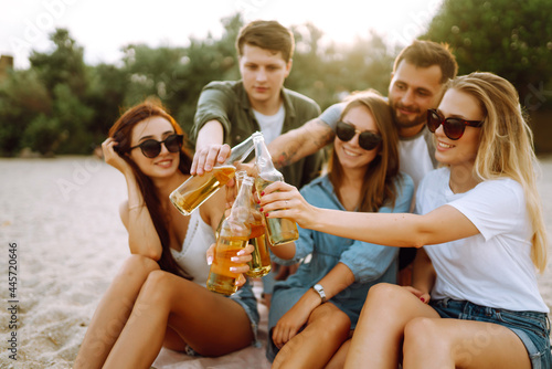 Group of friends cheers and drink beers on the beach. Young  friends sitting together sitting near the sea and enjoy summer party. Beach holiday and summer vacation concept. © maxbelchenko