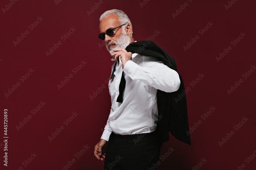 Adult man in white shirt and black sunglasses poses on burgundy background. Charming guy with grey beard in glasses and fashionable suit posing.. Adult man in white shirt and black sunglasses poses on burgundy background. Charming guy with grey beard in glasses and fashionable suit posing..