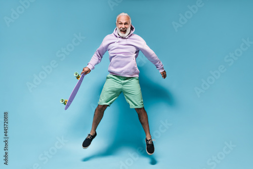 Cheerful man in hoodie jumping with skateboard on blue background. Happy bearded guy in purple clothes and shirt jumping..