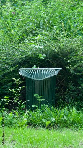 Green trash can in a public park by a bush