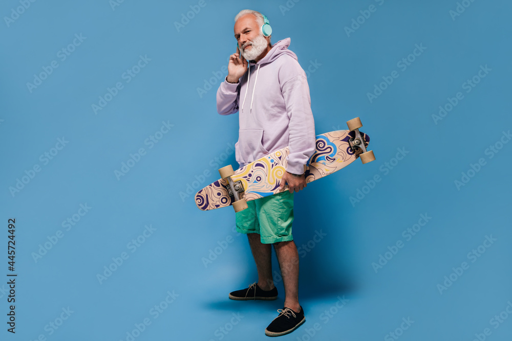Handsome adult man in lilac hoodie and shorts listening to music in headphones and holding skate board on isolated background.. Handsome adult man in lilac hoodie and shorts listening to music in headphones and holding skate board on isolated background..