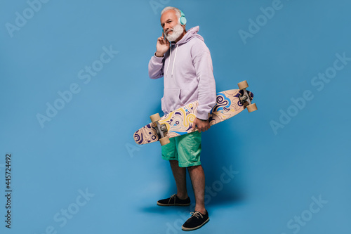 Handsome adult man in lilac hoodie and shorts listening to music in headphones and holding skate board on isolated background..