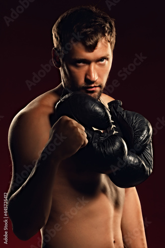 Portrait of a confident fighter on a dark background with a straight and bold look. Silhouette of a boxer with gloves around his neck. Caucasian fighter with a naked torso. © SerPak