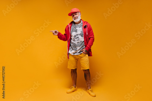 Man in red jacket and orange shorts pointing to place for text. White bearded guy with tattoos in modern bright clothes smiling..