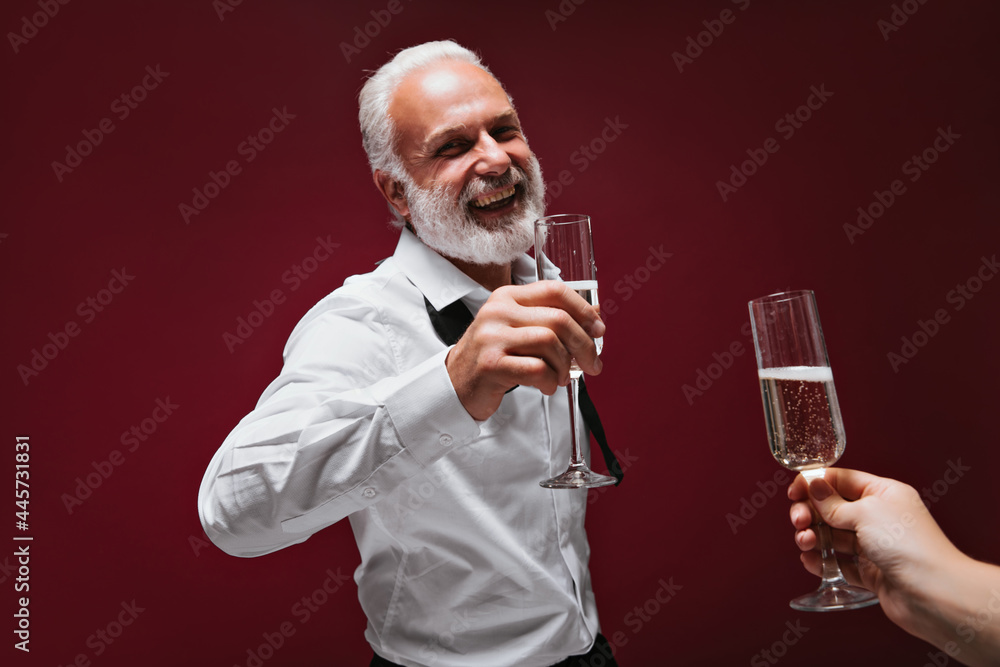 Joyful man in shirt holds champagne glases. Happy guy with beard in white modern clothes laughts on burgundy background.. Joyful man in shirt holds champagne glases. Happy guy with beard in white modern clothes laughts on burgundy background..