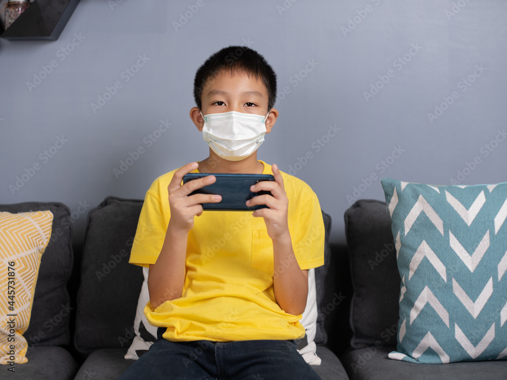 Happy asian boy wearing face masks sit on sofa at home holding phone looking at screen waving hand video calling distance friend online in mobile.