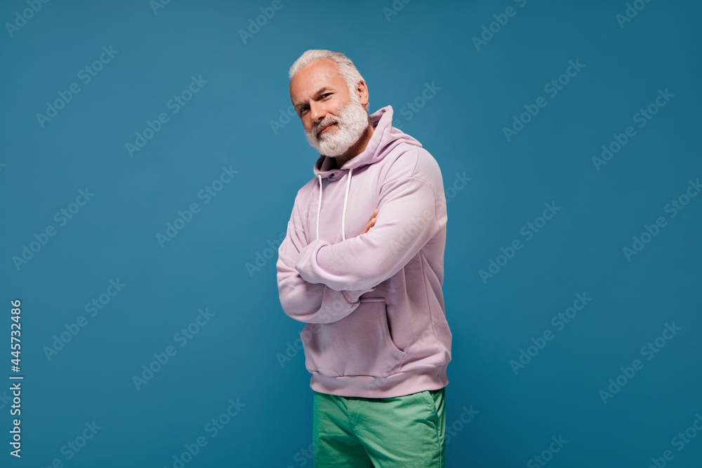 Fototapeta premium Portrait of man in sport style hoodie on blue background. Cool adult guy with grey hair in lilac sweatshirt and green pants posing..