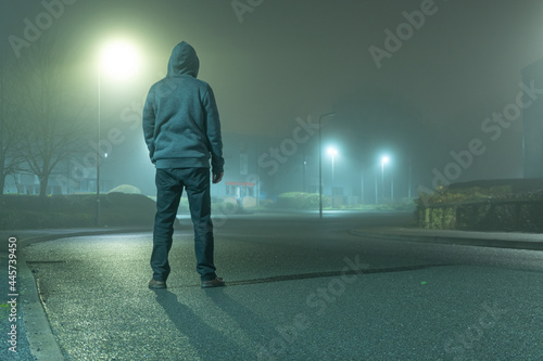 A mysterious hooded figure with back to camera, standing on a road in a light industrial urban area. On a moody, foogy, winters night photo