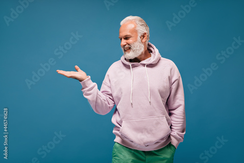 Happy man in hoodie smiling and pointing to place for text. Cool guy in good mood with beard in modern outfit posing on blue background..