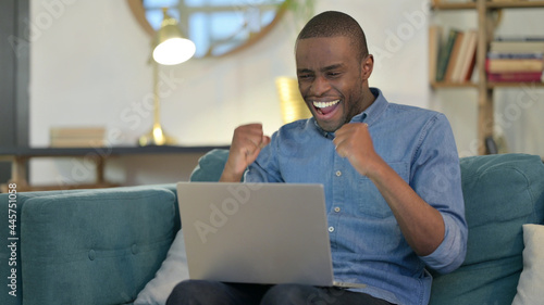 Successful Young African Man with Laptop Celebrating on Sofa 