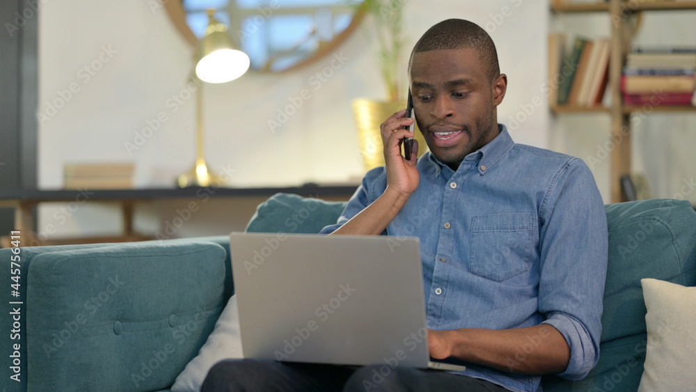 Young African Man with Laptop Talking on Smartphone on Sofa Young African Man with Laptop Talking on Smartphone on Sofa