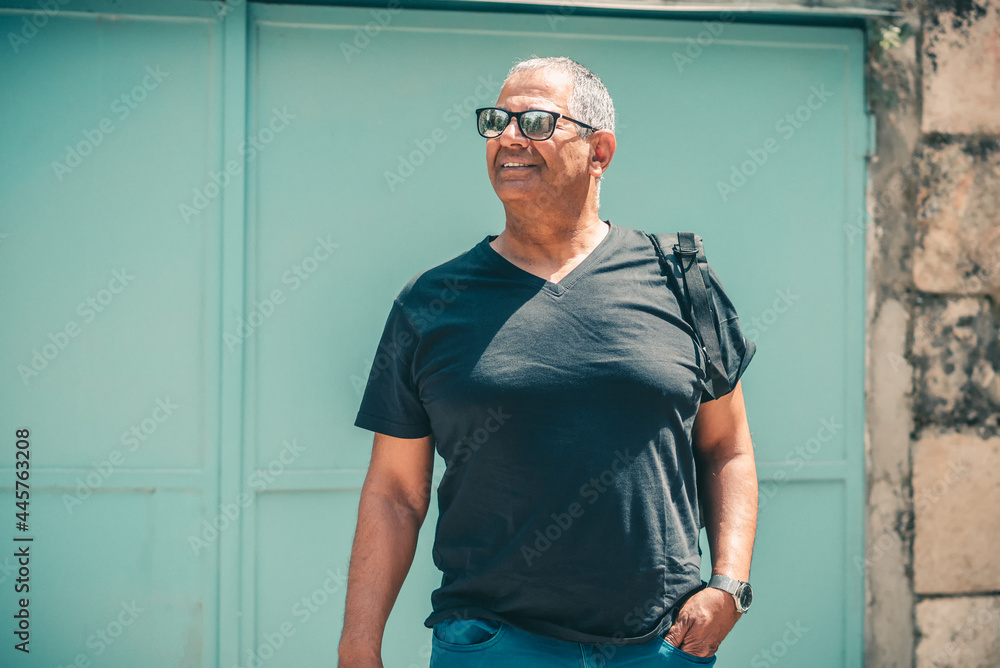 Smiling mature man standing in front of an old building holding his black backpack, looking away to side. Senior lifestyle travel. Smiling mature man standing in front of an old building holding his black backpack, looking away to side. Senior lifestyle travel.