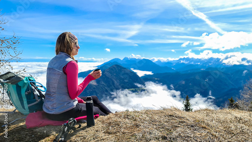 A woman in hiking outfit sipping a tea from thermos with the panoramic view from the top of Alpine peak in Austria. The area is shrouded in thick clouds. A few peaks popping out from the clouds. Happy