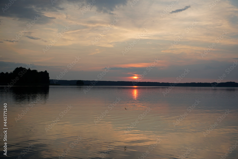 Evening on the lake in summer in July. The sun and sky are reflected on the calm surface of the water. Soft light and bright colors. Sunset and pacification