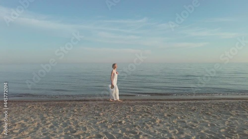 Woman wearing long white dress walk along beachof the calm sea barefoot at early morning, slow motion photo