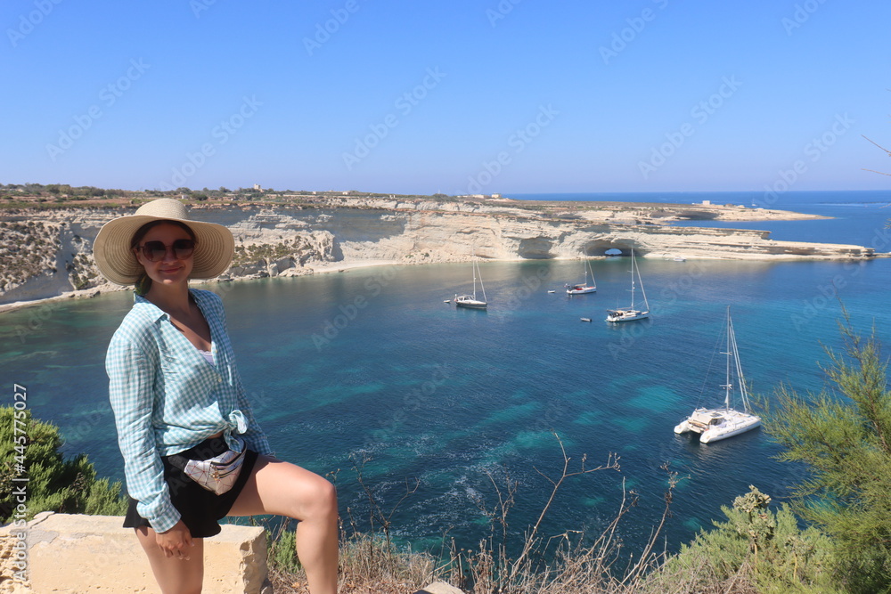Girl stand on the clif with hat on the head and sunglasses smiling in camera see backstage with beautiful azure water bay in Marsaxlokk Malta