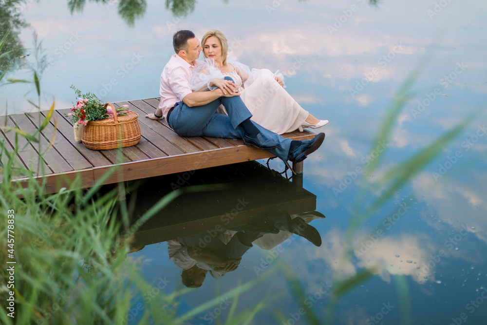 Happy romantic mature couple sitting on a wooden bridge near lake or pond outdoors, hugging and enjoying their date. Wedding anniversary celebration Happy romantic mature couple sitting on a wooden bridge near lake or pond outdoors, hugging and enjoying their date. Wedding anniversary celebration