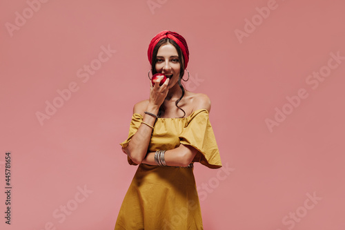 Modern woman in red headband, round earrings and yellow dress eating apple and looking into camera on isolated background..