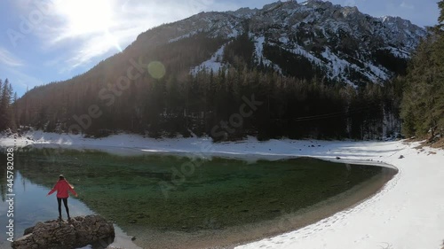 A woman in pink jacket standing on the stone by the shore of Green Lake in Austria in winter. The lake is surrounded by snow. There is a dense forest around the lake. Sunny and bright day. Relaxation
 photo