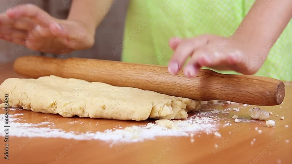 girl in an apron in the kitchen rolls out the dough with rolling pin for making cookies on wooden table, close-up girl in an apron in the kitchen rolls out the dough with rolling pin for making cookies on wooden table, close-up