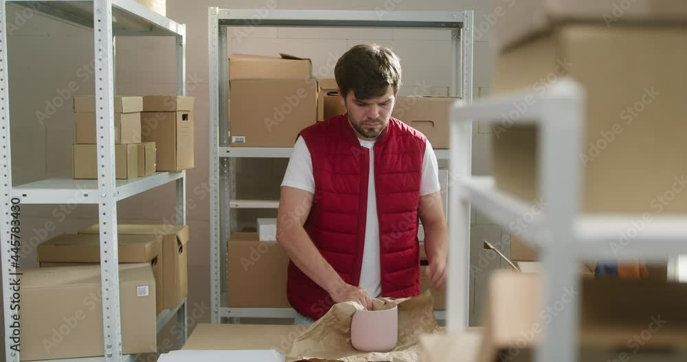 Man wearing red vest, employee of warehouse packing parcel into delivery box, closing and putting on shelf on the background of cardboard boxes. Logistics, delivering and storage concept. Man wearing red vest, employee of warehouse packing parcel into delivery box, closing and putting on shelf on the background of cardboard boxes. Logistics, delivering and storage concept.