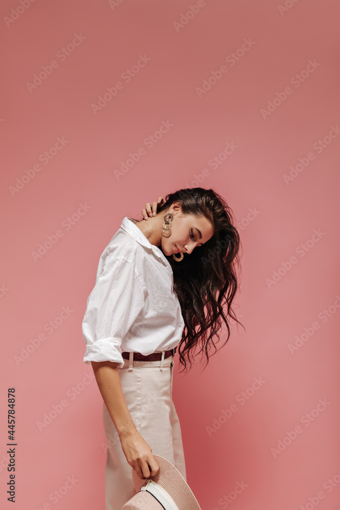 Modern wavy haired lady with cool accessories in wide light shirt and beige trousers holding hat and posing on isolated backdrop.. Modern wavy haired lady with cool accessories in wide light shirt and beige trousers holding hat and posing on isolated backdrop..