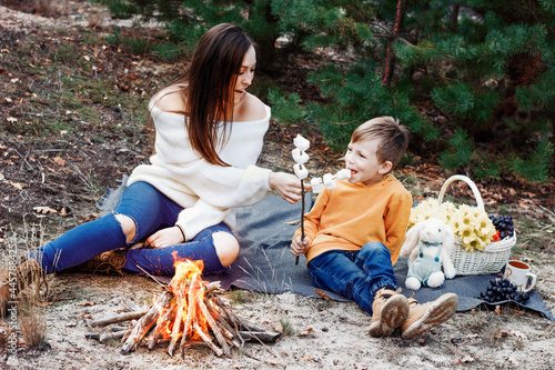 Young mother with little son roasting marshmallows over the fire. Happy family at the autumn picnic in the pine forest