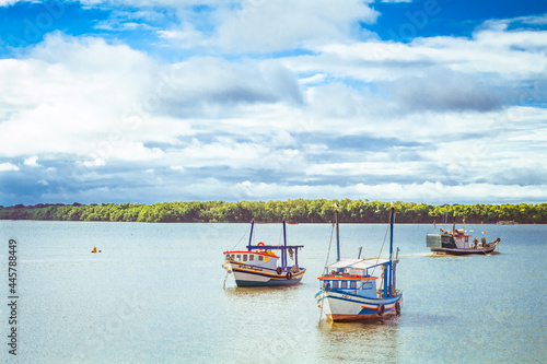 Three fishing boats in the sea with the shore in the background, trees and clouds in the blue sky. Cananéia, São Paulo, Brazil.
