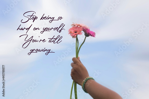 Inspirational motivational quote - Stop being so hard on yourself, you're still growing. With hand of young woman holding pink gerber flowers against summer sky background. Self love care concept. photo