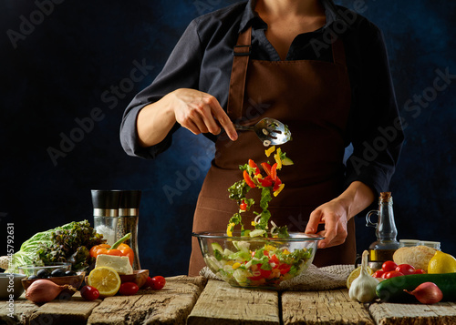 The cook mixes the prepared Greek salad in a large glass bowl with a spoon. Levitating lettuce ingredients. Many ingredients are on the table near the bowl. Wooden table. Dark blue background.