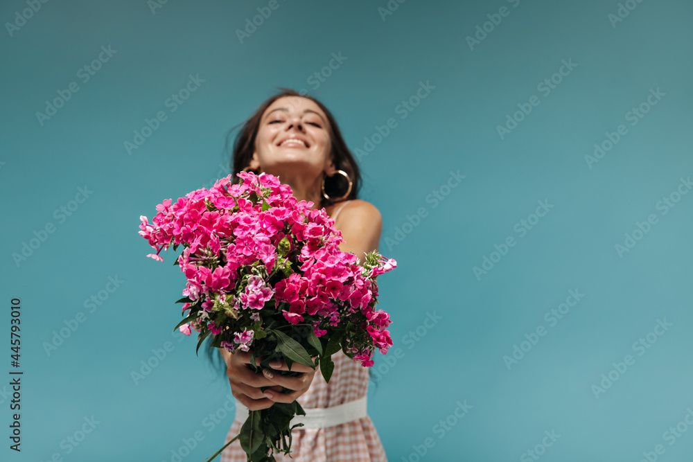 Joyful summer women with brunette hair in cool light sundress holding bright pink wildflowers and smiling on blue background.. Joyful summer women with brunette hair in cool light sundress holding bright pink wildflowers and smiling on blue background..