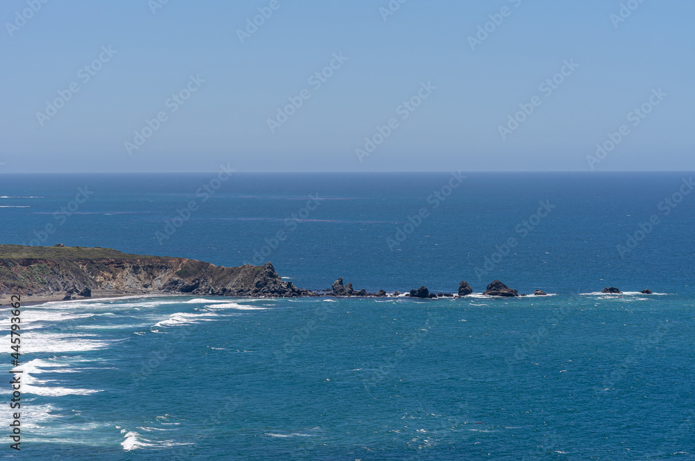 San Simeon, CA, USA - June 8, 2021: Pacific Ocean coastline. South of Ragged Point, black rocks form line into deep blue ocean extanding from black rock cliffs, under light blue sky.