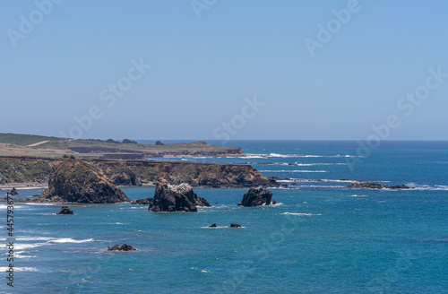 San Simeon, CA, USA - June 8, 2021: Pacific Ocean coastline North of town. Landscape with Black cliffs and rocks in the deep blue water under light blue sky. White surf adds color.