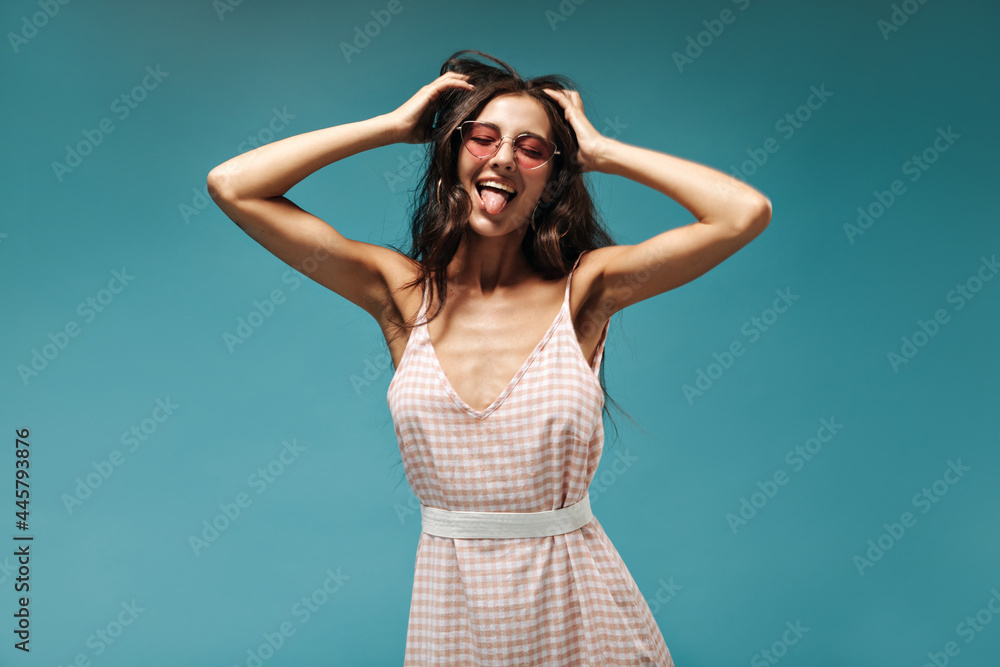 Funny stylish girl with curly dark hair in pink cpp sunglasses and plaid fashionable clothes showing her tongue with closed eyes.. Funny stylish girl with curly dark hair in pink cpp sunglasses and plaid fashionable clothes showing her tongue with closed eyes..