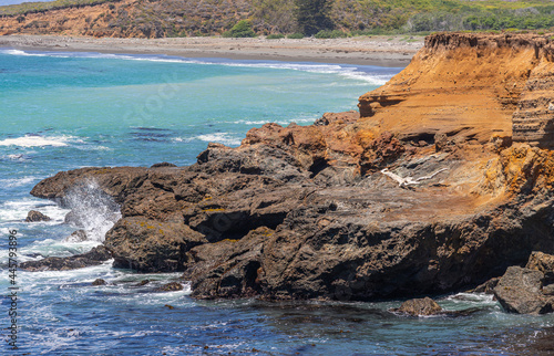 San Simeon, CA, USA - June 8, 2021: Pacific Ocean coastline north of town. White driftwood on deep brown rocky shoreline circled by blue-azure water.