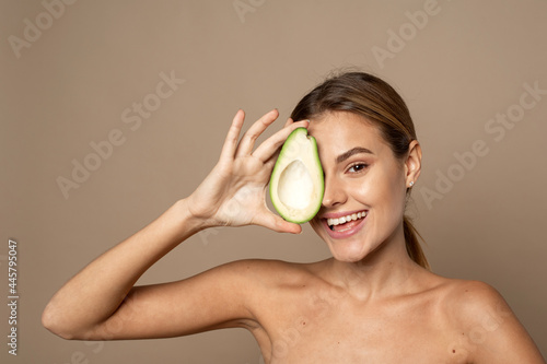 Happy smiling young woman holding avocado half on a beige background. Natural beauty concept photo