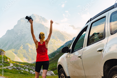 Caucasian young woman with arms raised and holding a photo camera near her off-road car during an adventure trip up the mountain.