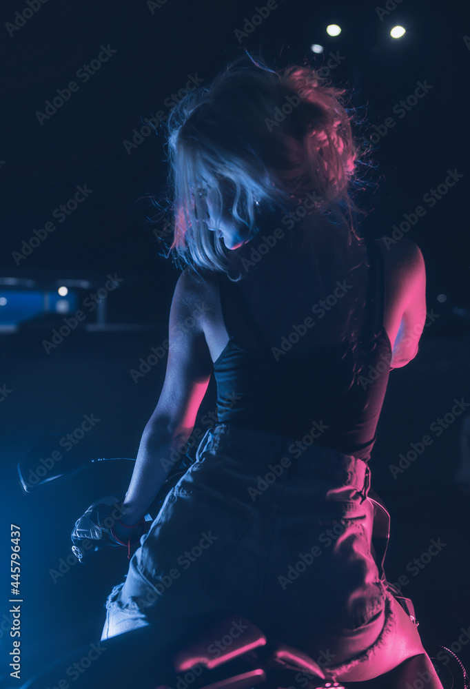 portrait of a girl in the rays of neon light on a motorbike at night in an empty parking lot