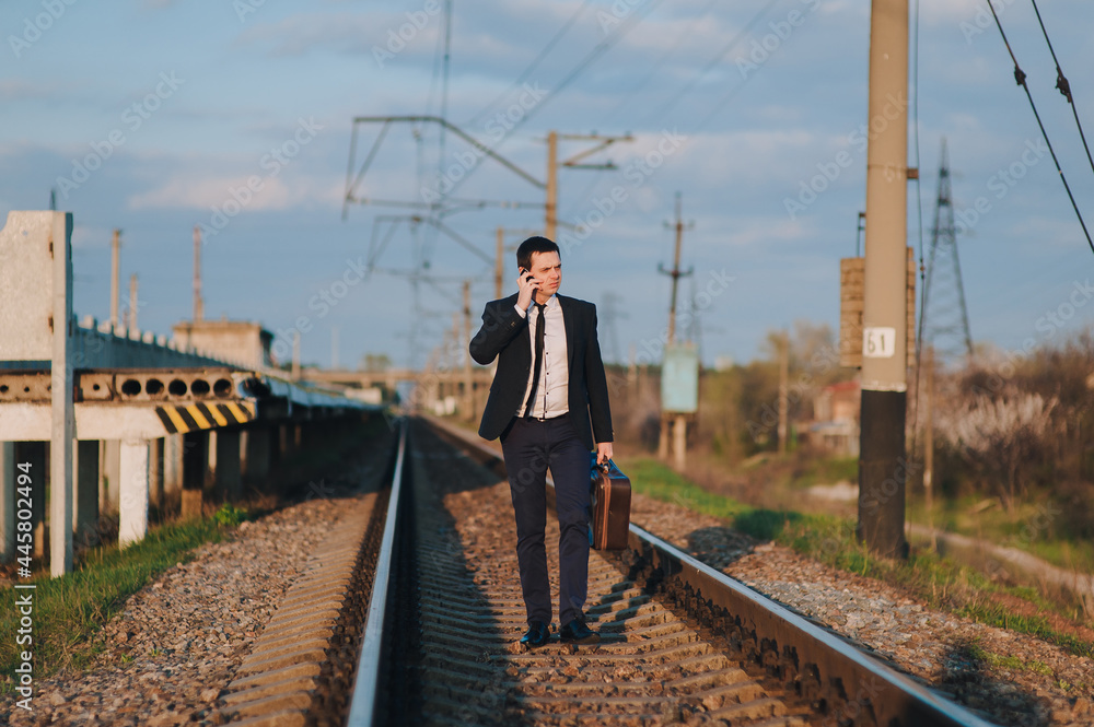 Stylish young male businessman in a suit with a suitcase is walking along the tracks from the train and talking on the phone. Business walk and serious conversation. Late for the train.